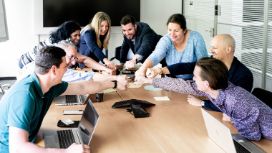 Ein Team sitzt mit Laptops an einem Tisch und beugt sich gemeinsam für einen Handschlag nach vorne.