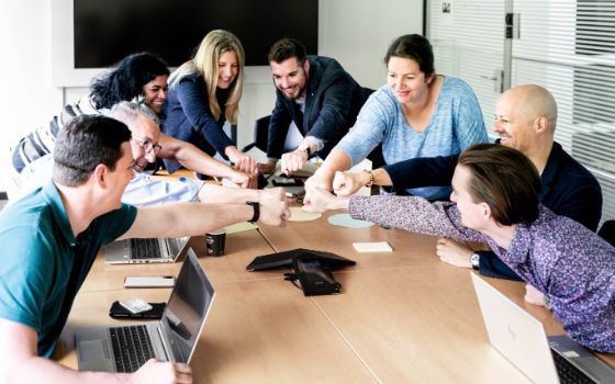 Ein Team sitzt mit Laptops an einem Tisch und beugt sich gemeinsam für einen Handschlag nach vorne.